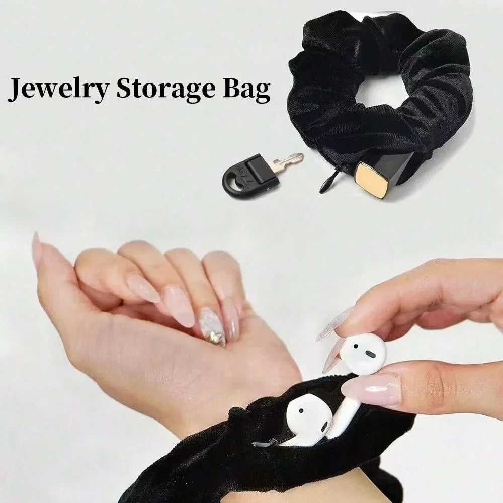 Black velvet hair scrunchie with a zipper pocket, worn on a wrist and holding wireless earbuds.