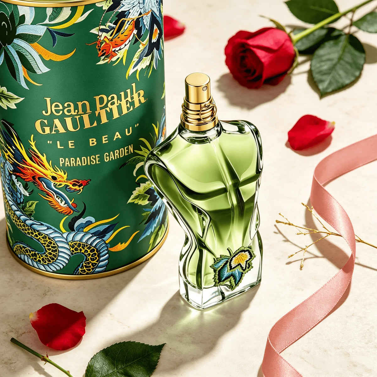 Green glass torso shaped perfume bottle with a vine leaf detail next to its decorated tin, red rose petals, and a pink ribbon