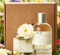A glass bottle of scented perfume spray with a label, presented with a white rose in front of a cardboard box.