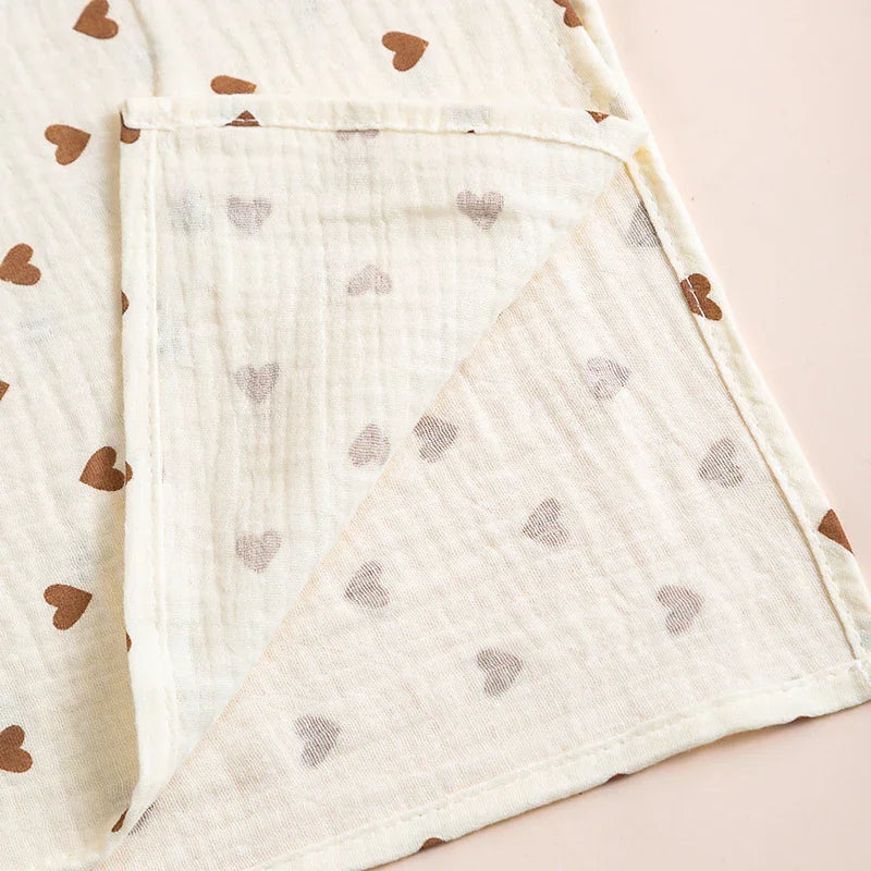 Close-up corner view of a soft, cream-colored cotton muslin baby blanket with a brown heart pattern, showing the hemmed edge.
