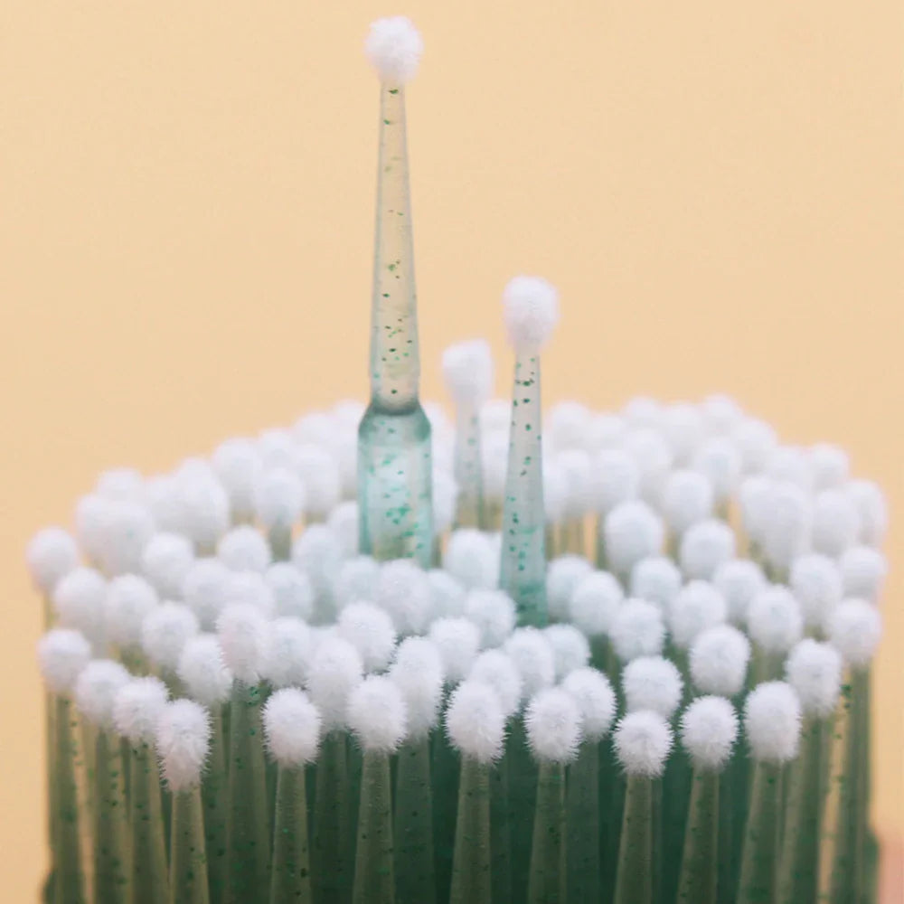 Close-up view of several green disposable microbrushes with white fuzzy tips for applying lash extension products.
