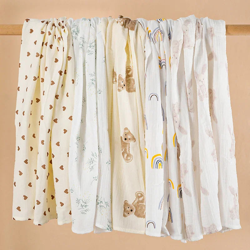 Set of several cotton muslin baby blankets in various patterns including hearts, teddy bears, and rainbows, draped over a