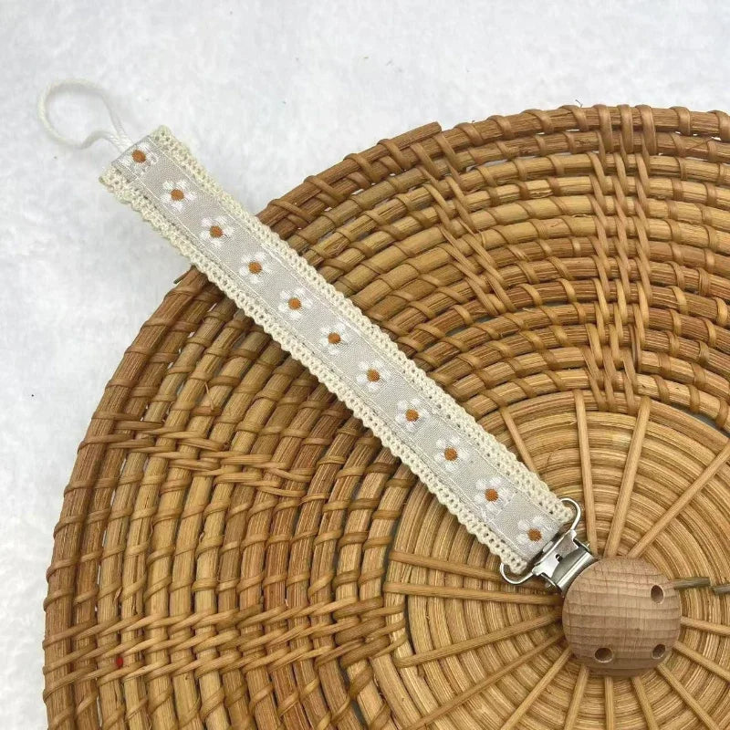 A single fabric pacifier chain with a small daisy flower pattern and a wooden clip on a woven tray.