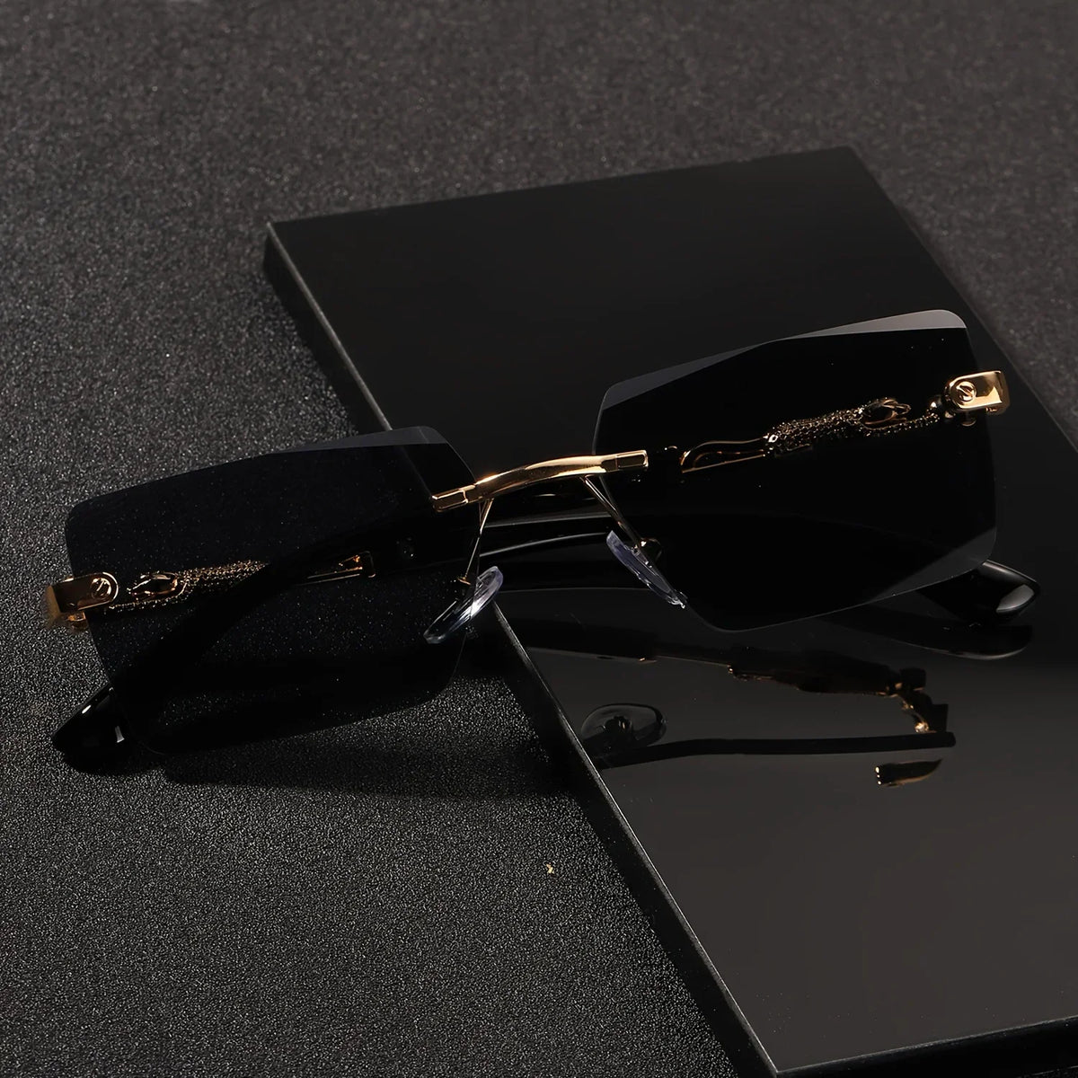 Rimless rectangular sunglasses with black lenses and gold panther-style metal temples on a black display base