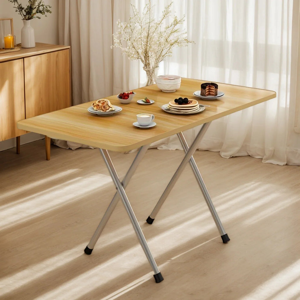A small light-colored wood folding table with metal legs, set with plates of food, near white curtains.