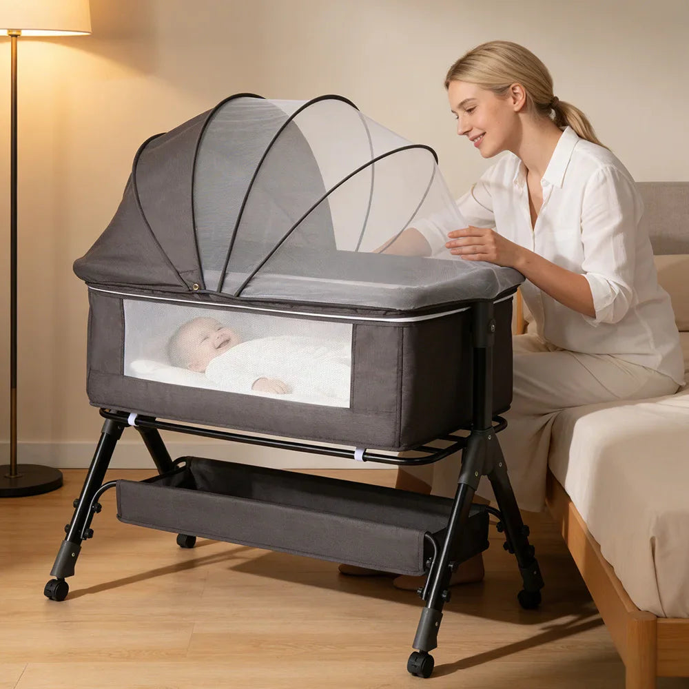 A grey baby bedside sleeper bassinet with a mosquito net and an infant resting inside, with a woman looking on.