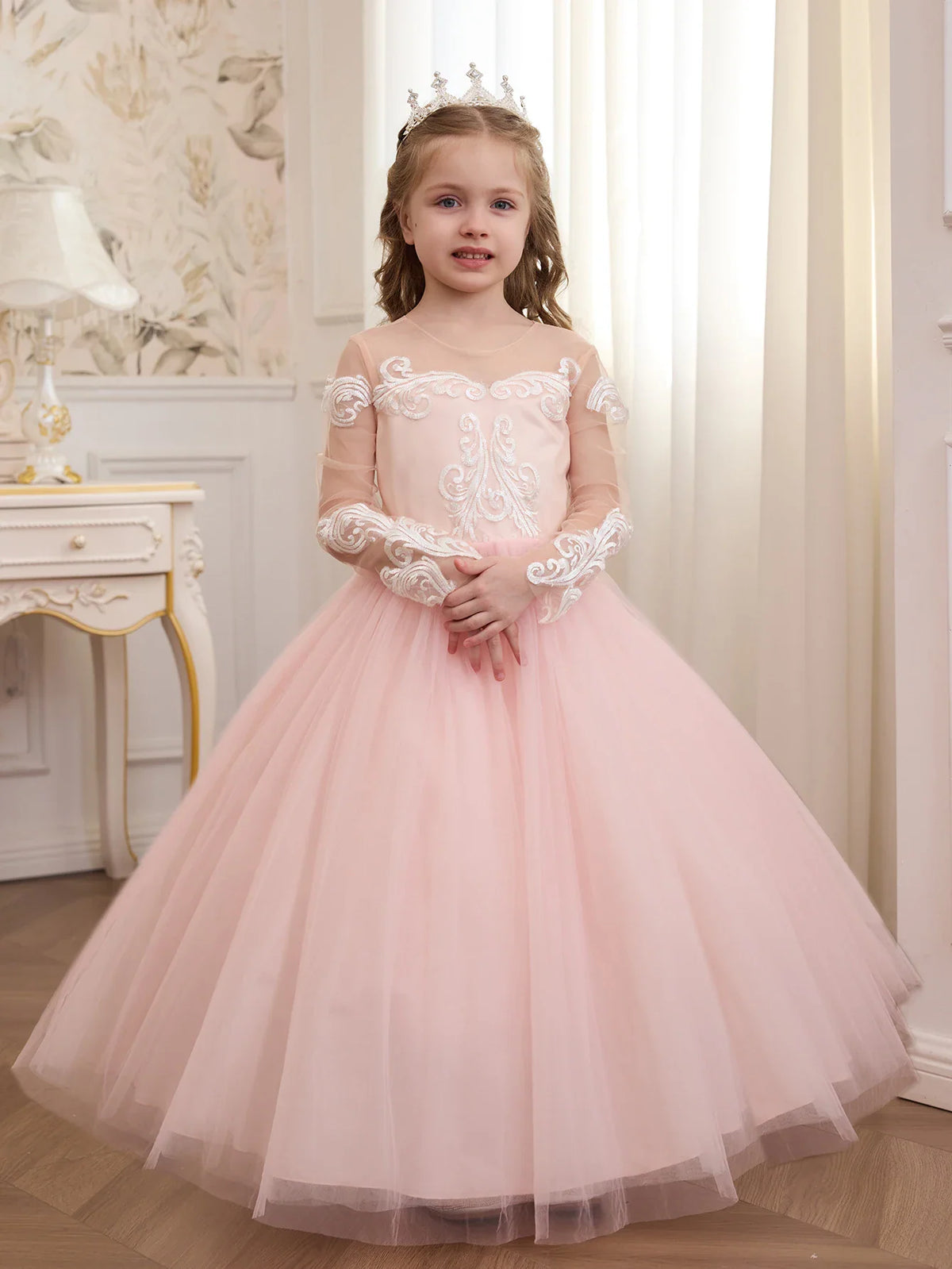 A young girl in a pale pink floor-length gown with white floral lace embroidery on the bodice and long sleeves.
