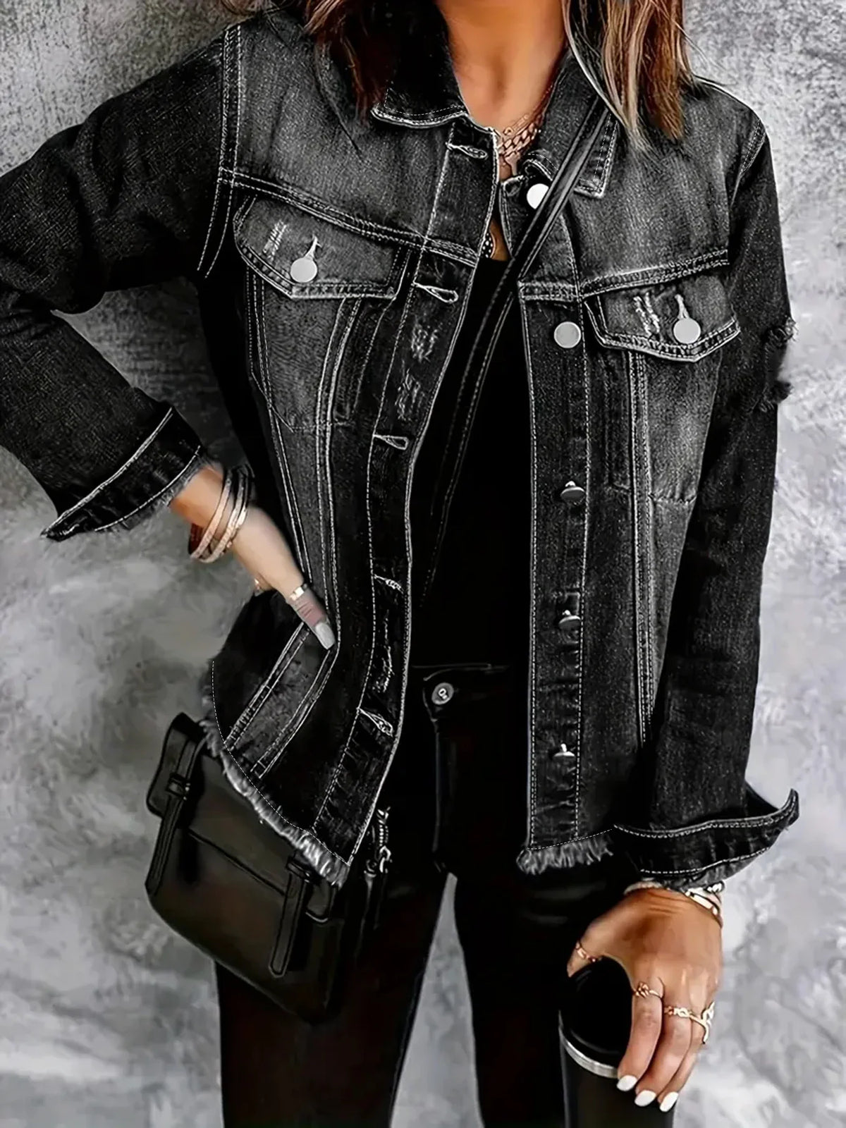 Woman wearing a dark grey distressed denim jacket over a black shirt, posing with a black shoulder bag