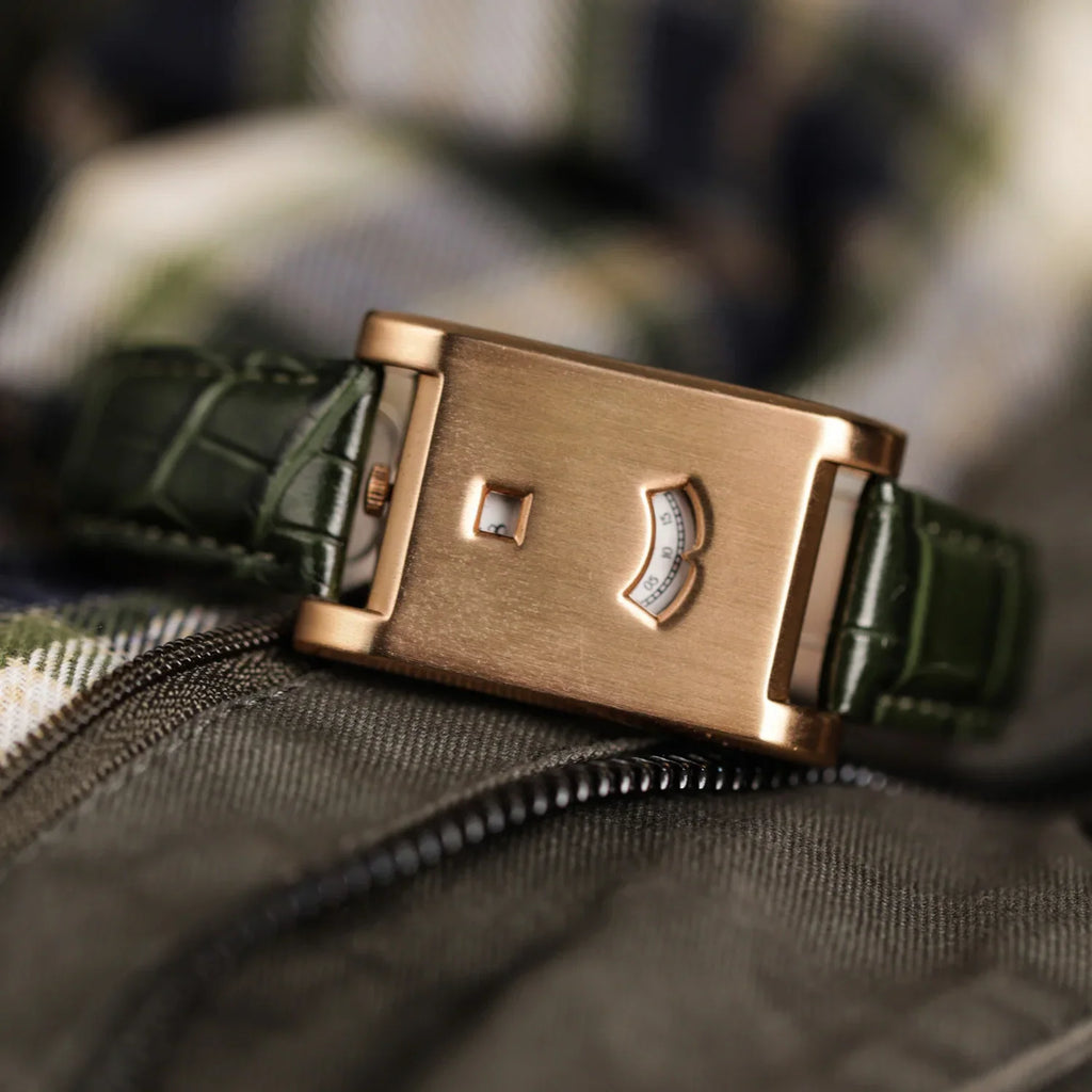 A rectangular rose gold watch with a green alligator strap set on a plaid and dark fabric surface.