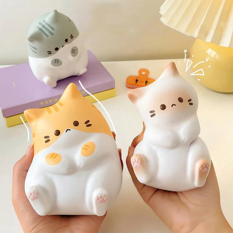 Hands holding a yellow and white cartoon cat stress relief toy with other toy cats and books in the background.
