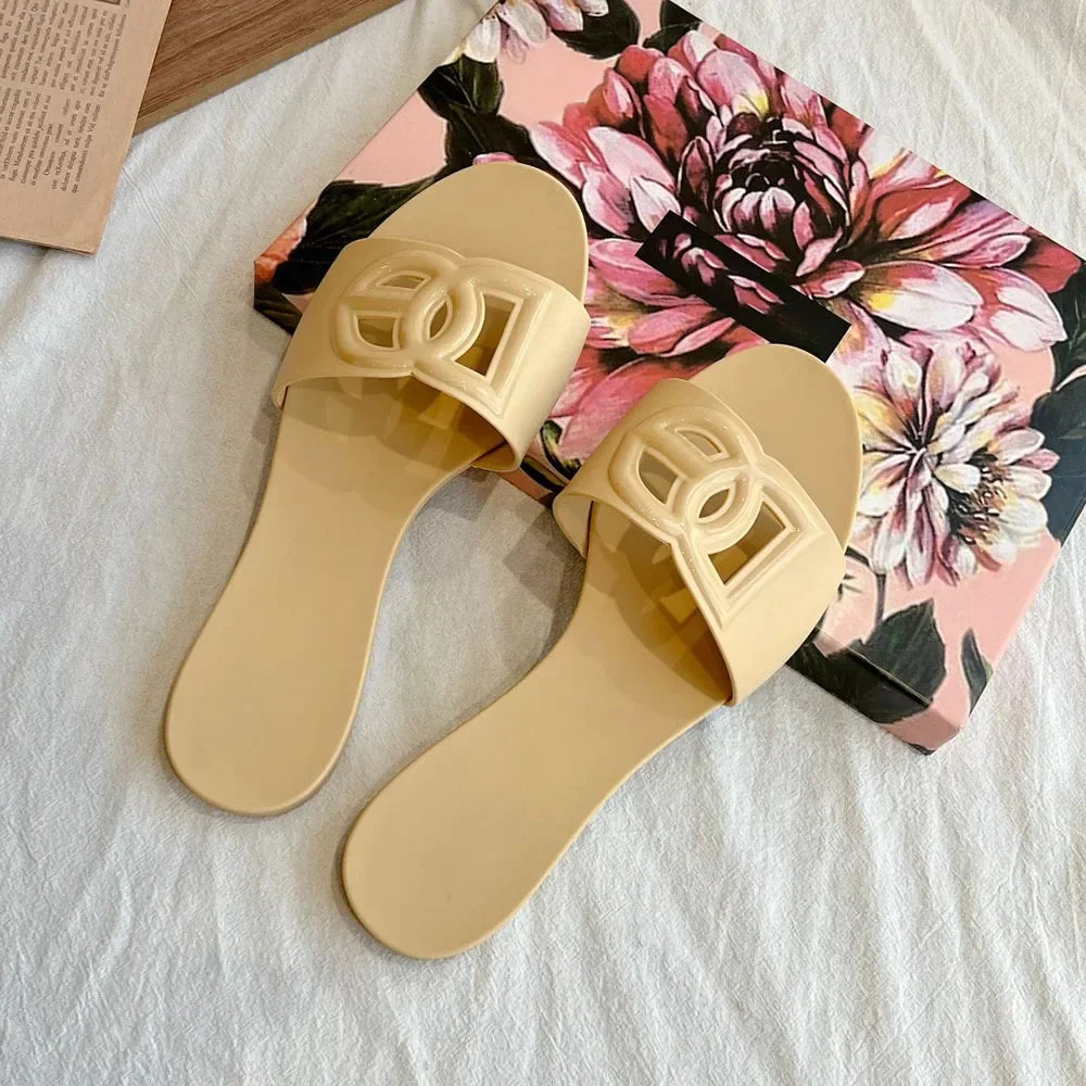 Pair of beige jelly slides with a decorative cutout letter emblem on the strap, resting on a floral box.