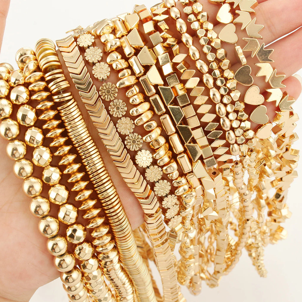 A hand holding multiple strands of gold-tone jewelry beads in various geometric shapes including round, cube, and heart.