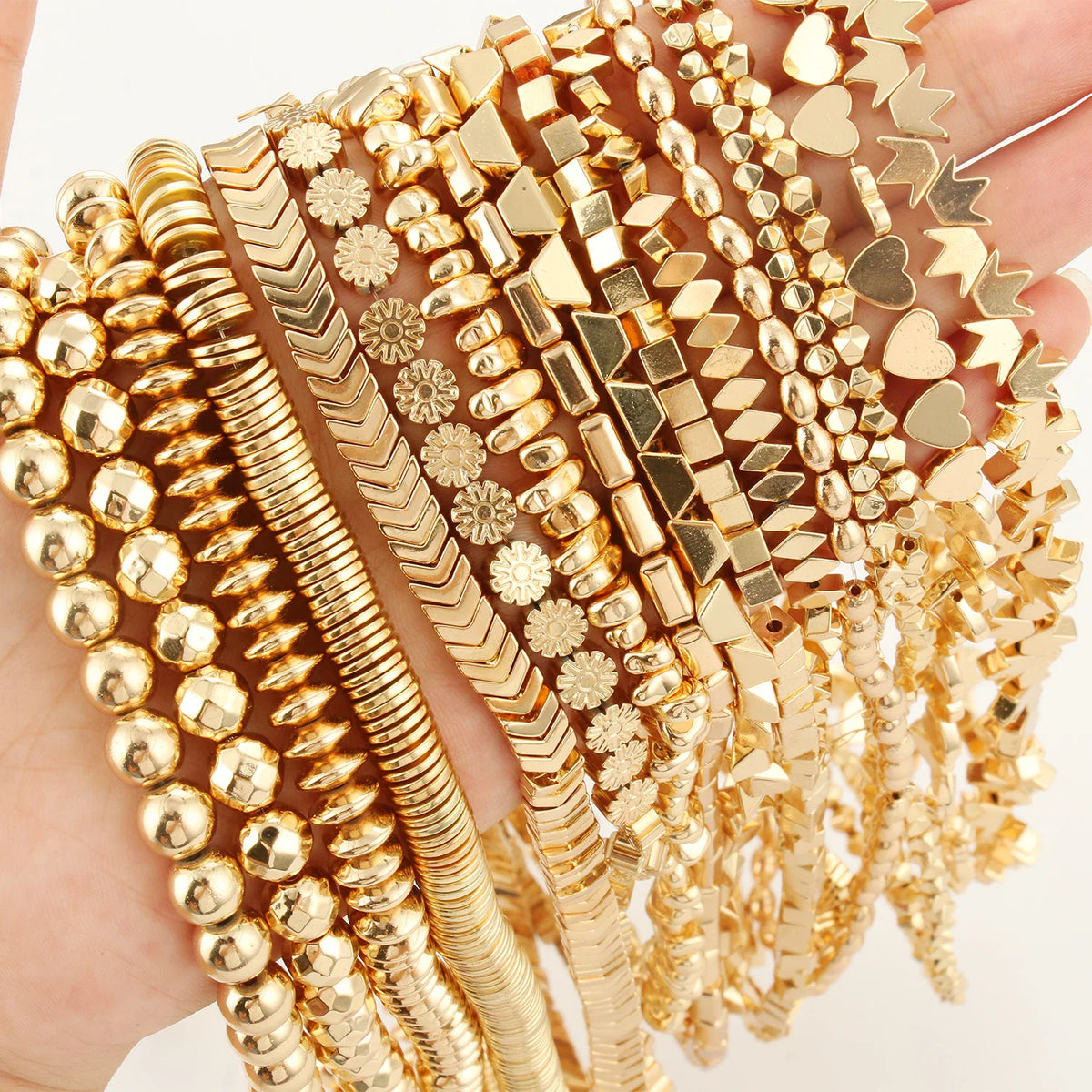 A hand holding multiple strands of gold-tone jewelry beads in various geometric shapes including round, cube, and heart.