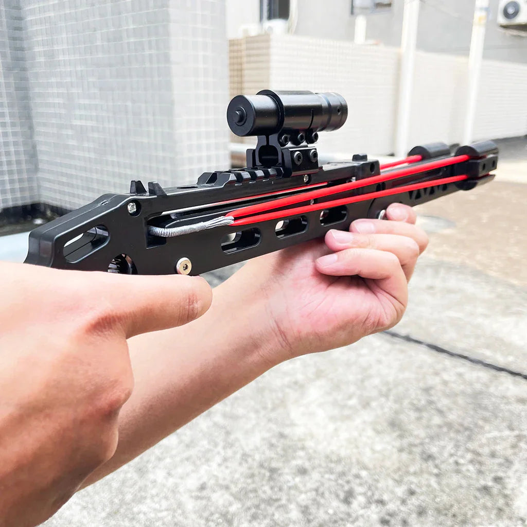 Person aiming a black foldable slingshot with red elastic bands and tactical sight
