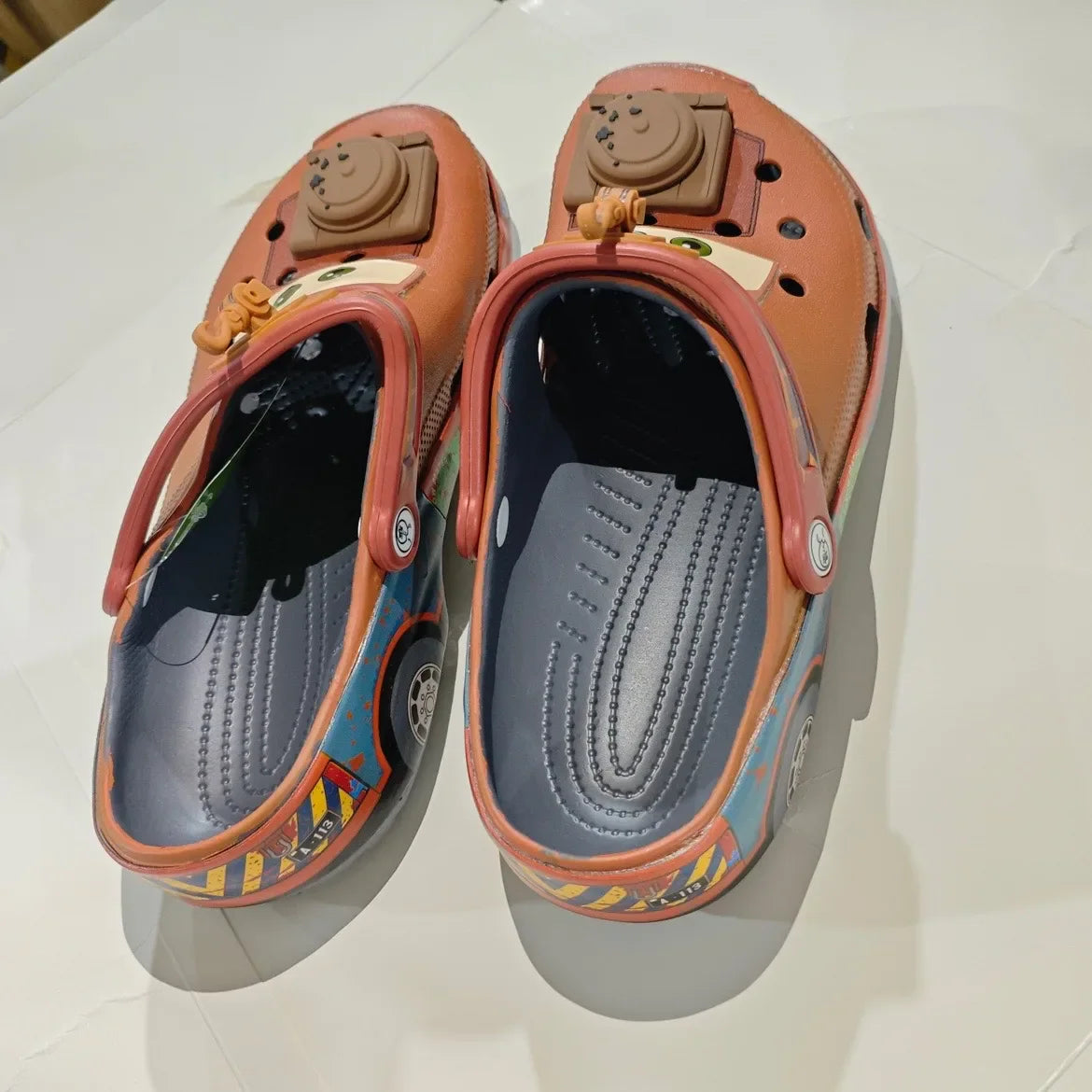 Angled rear view of brown cartoon tow truck themed clogs showing inner soles and tire patterns.