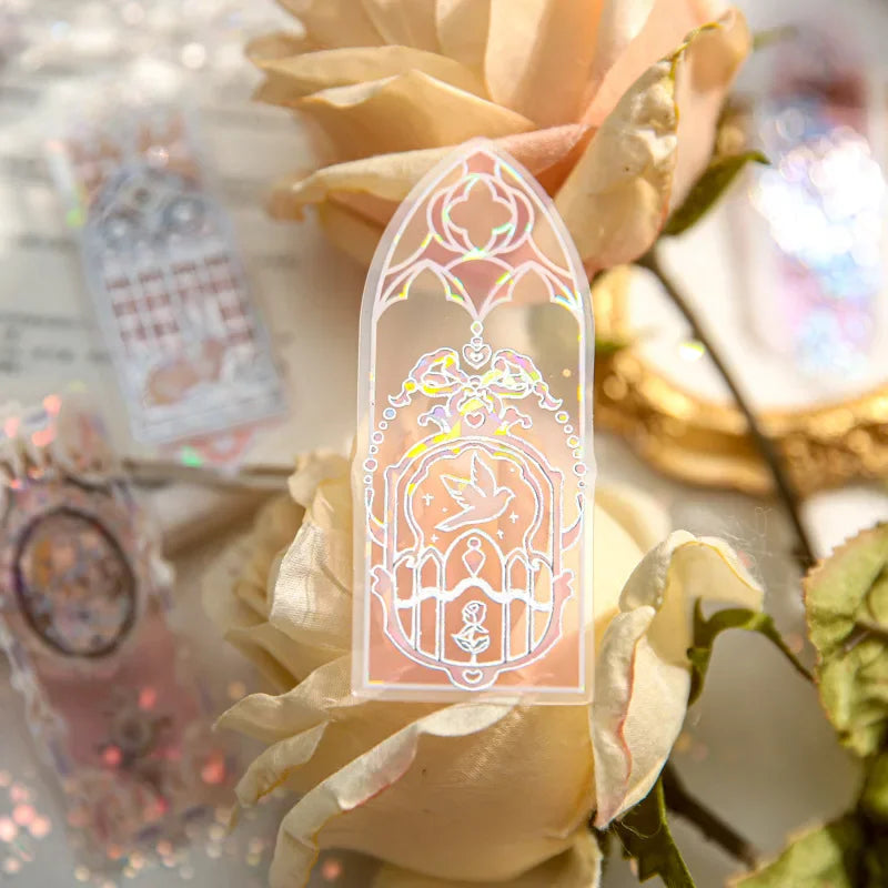 A single transparent holographic bookmark with a dove and rose motif, held in front of white roses.