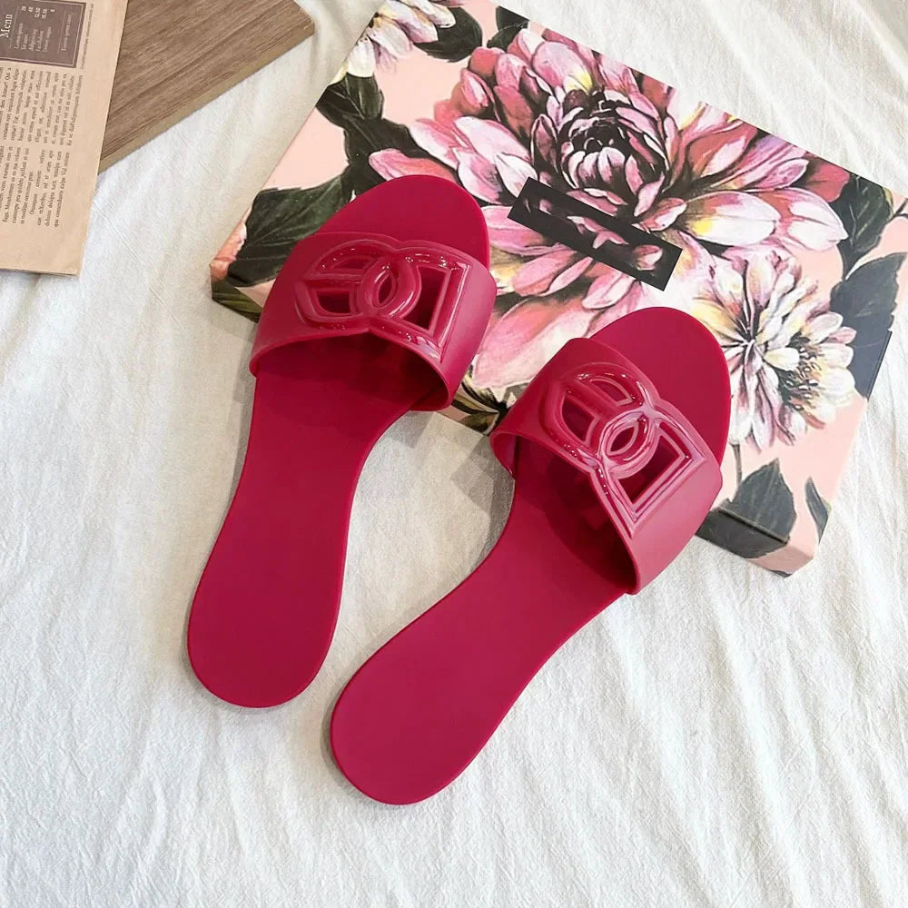 Pair of dark pink jelly slides with a decorative cutout letter emblem on the strap, resting on a floral box.