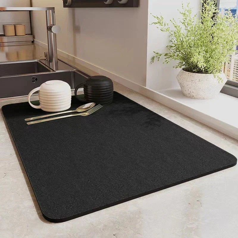 Black absorbent kitchen drying mat on a countertop with cups and cutlery.