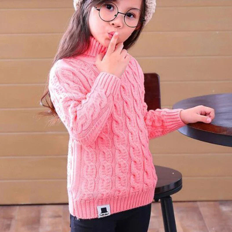 A young girl wearing a pink cable-knit turtleneck sweater with a mustache logo patch.