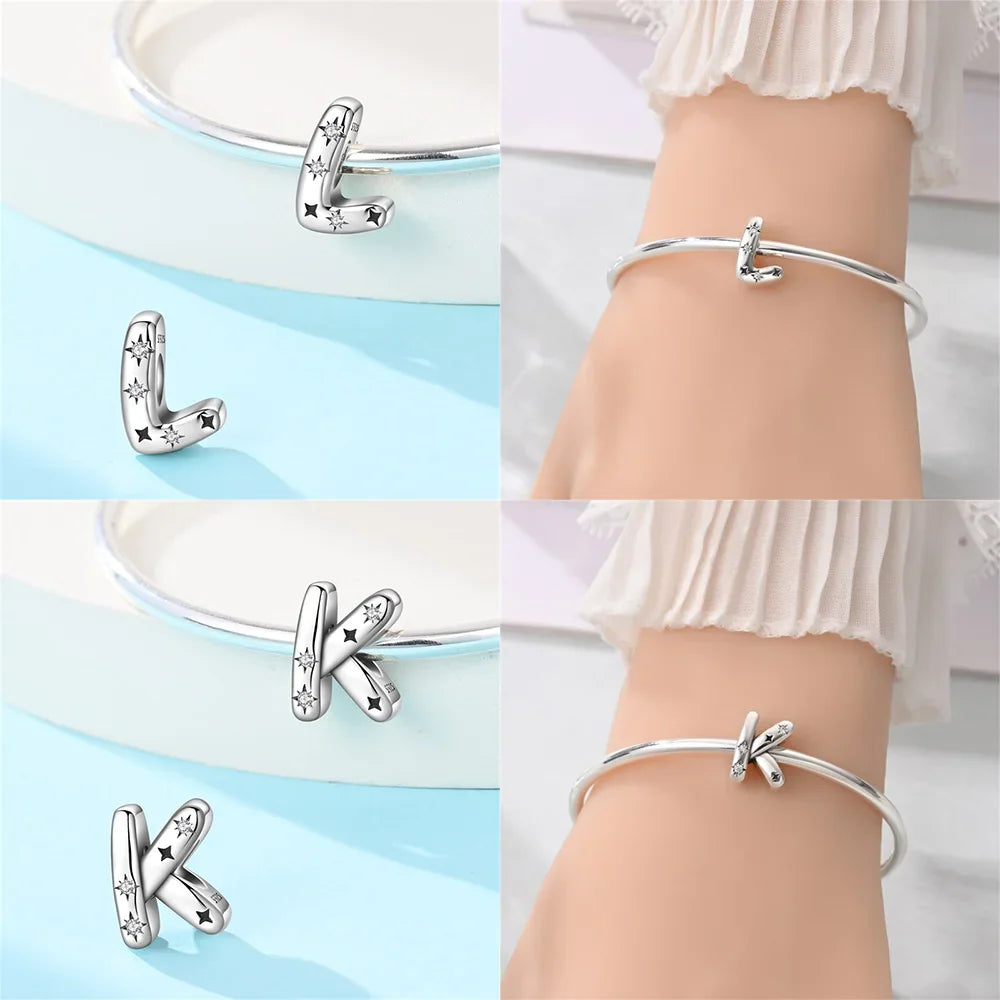 Sterling silver alphabet charms L and K, shown on a light blue display and worn on a bracelet on a wrist.