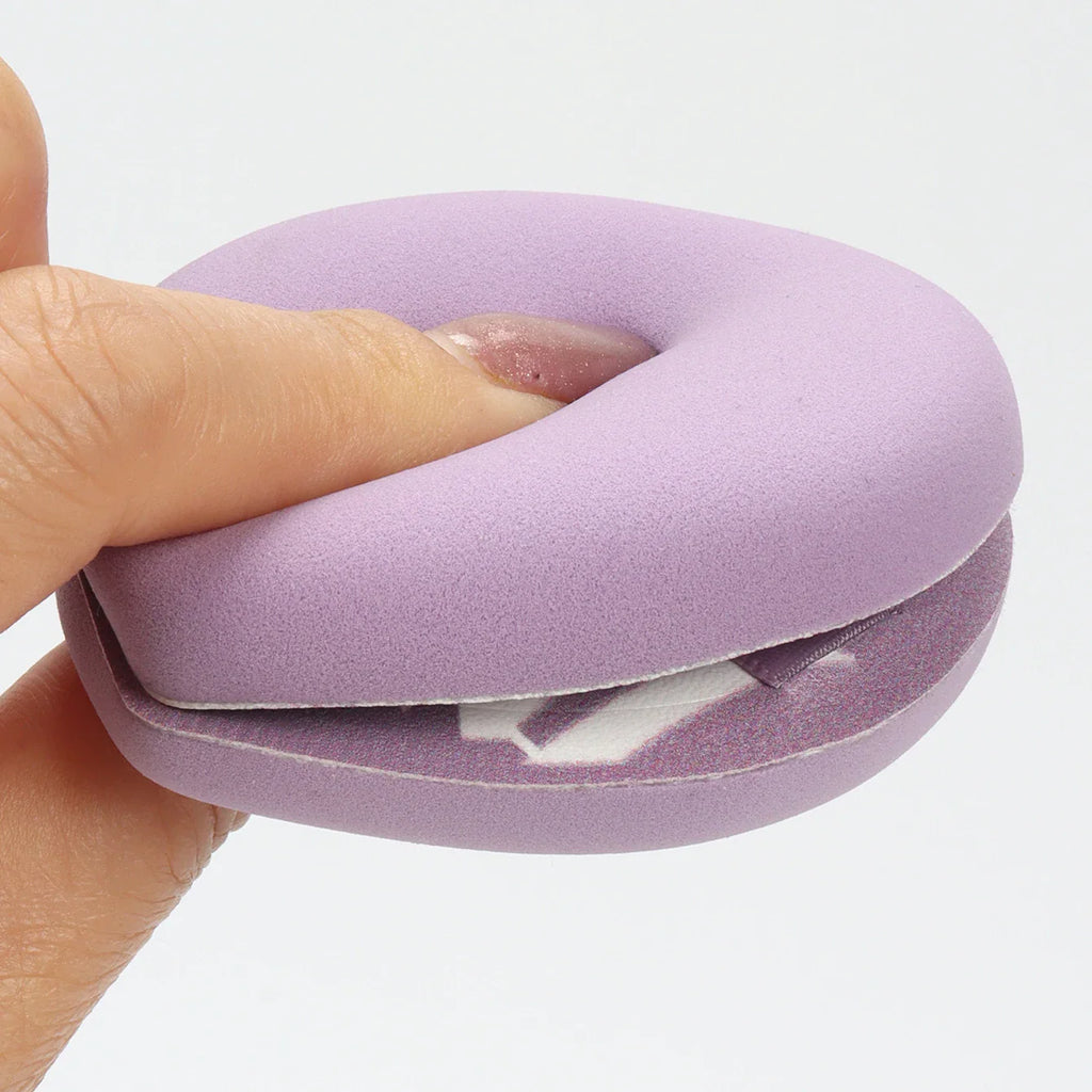 A hand squeezing a soft purple circular makeup sponge to demonstrate elasticity