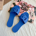 Pair of bright blue jelly slides with a decorative cutout letter emblem on the strap, resting on a floral box.
