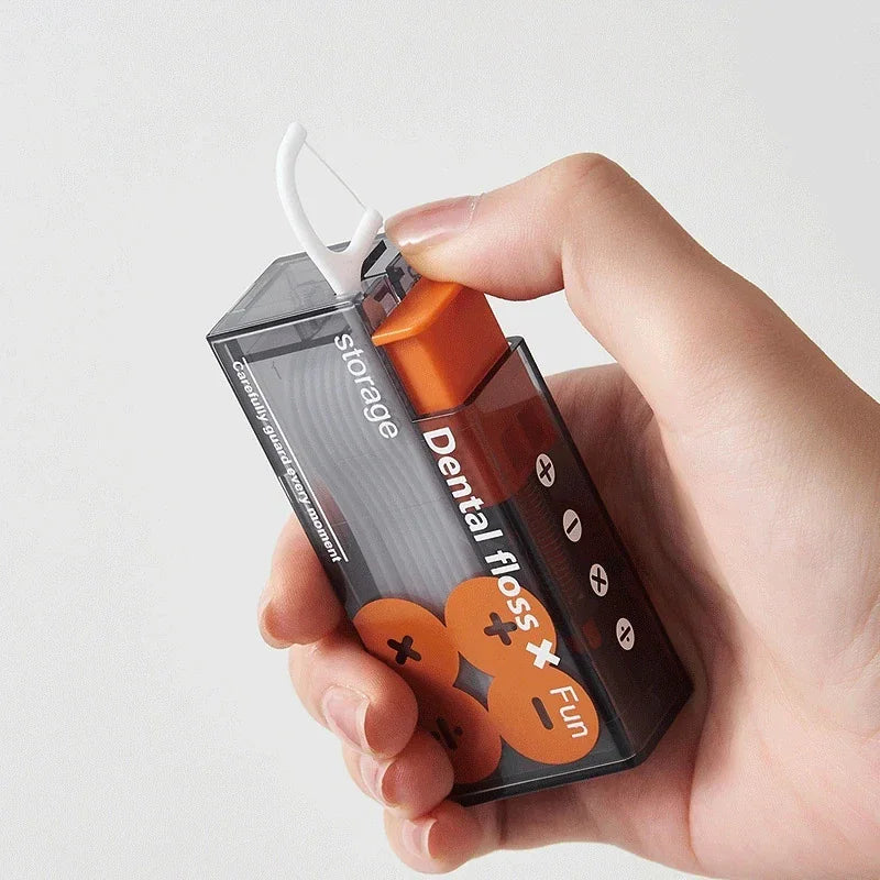 A hand holding a portable rectangular dental floss dispenser in black and orange with automatic ejection.
