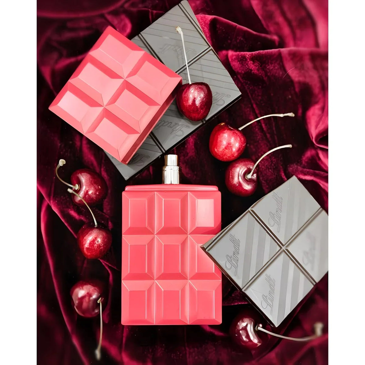 A perfume bottle shaped like a pink chocolate bar surrounded by chocolate squares, cherries, and red fabric.