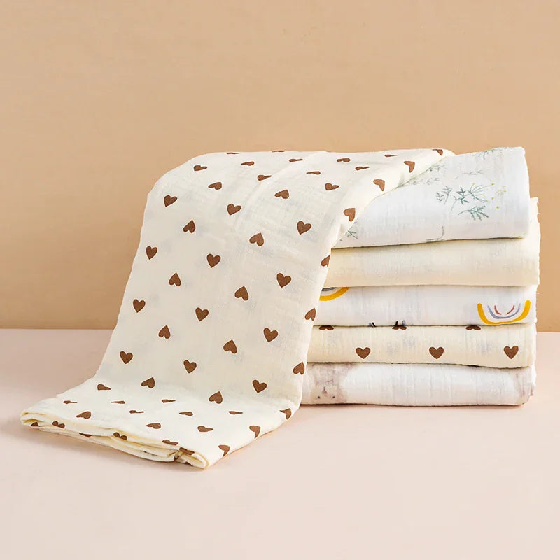A stack of folded cotton muslin baby blankets featuring different patterns, including hearts, bears, and botanical prints.