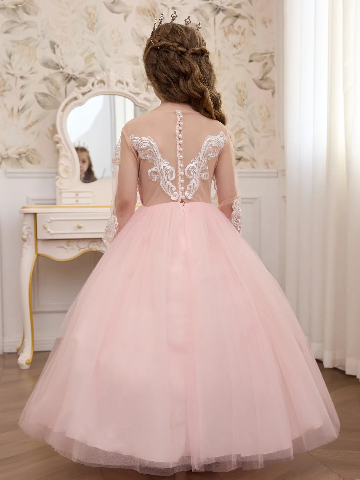 The back view of a young girl in a pink tulle dress featuring a sheer bodice, white lace detailing, and pearl button