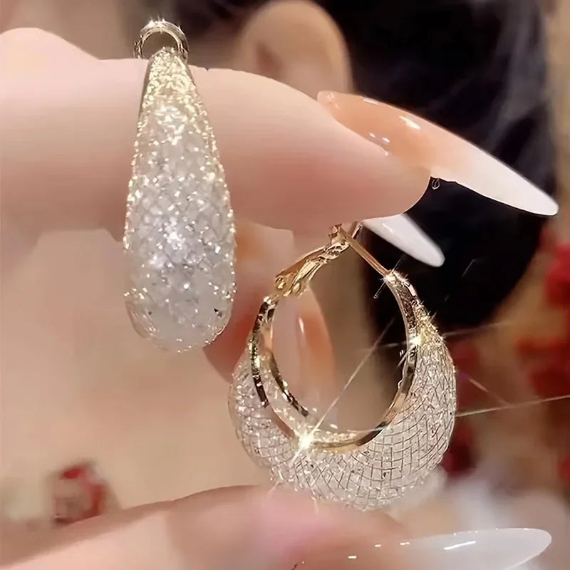 Shiny gold-tone hoop earrings with mesh texture and rhinestones held in fingers