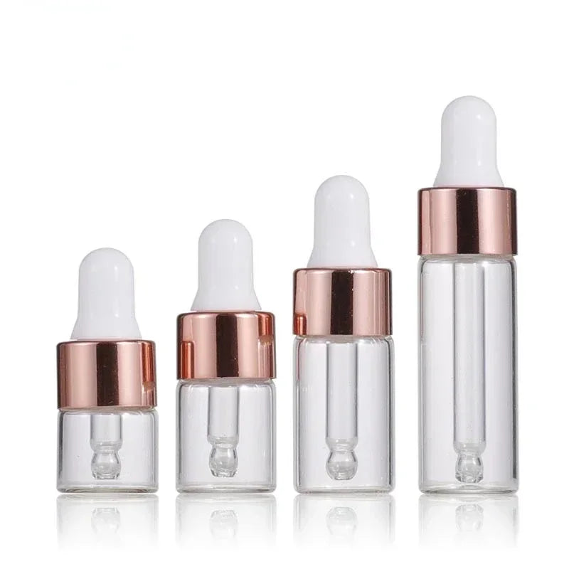 A lineup of four clear mini glass dropper bottles of varying sizes with rose gold caps and white silicone droppers.