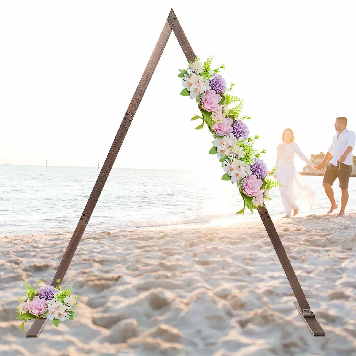 A rustic wooden triangular wedding arch decorated with flowers, positioned on a beach with a couple in the background.