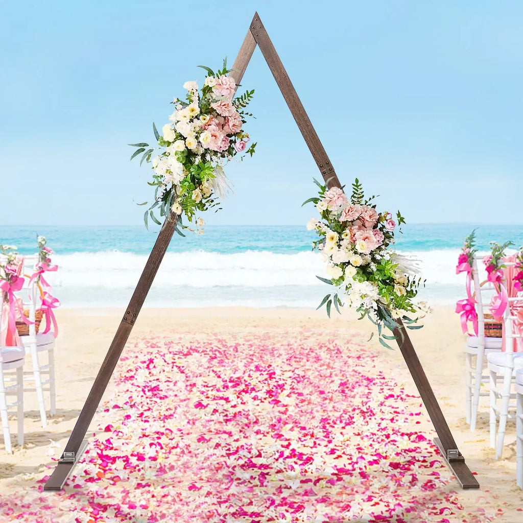 A rustic wooden triangular wedding arch decorated with artificial flowers on a beach with a pink flower petal aisle.