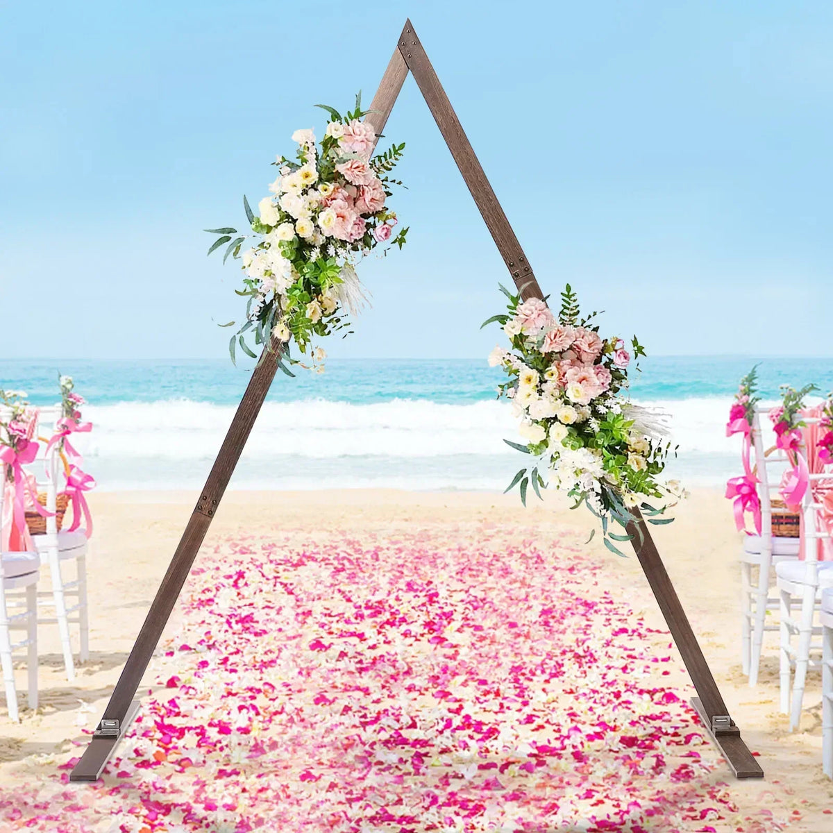 A rustic wooden triangular wedding arch decorated with artificial flowers on a beach with a pink flower petal aisle.