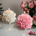 A white and a pink peony shaped candle burning on a table with a rose bouquet