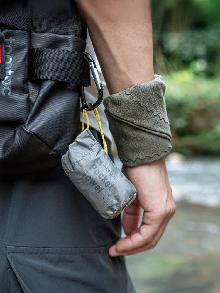 A hand holding a grey folded pocket towel with a carabiner attached to a backpack strap.