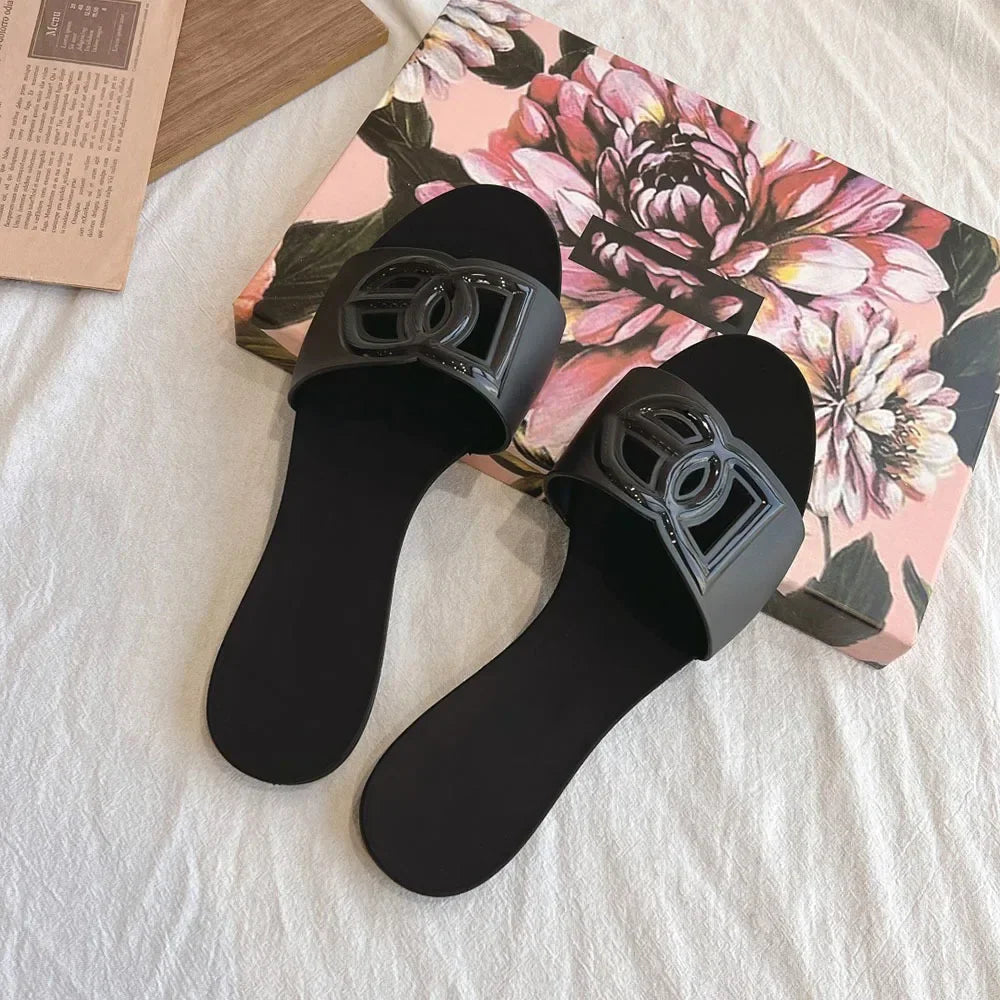 Pair of black jelly slides with a decorative cutout letter emblem on the strap, resting on a floral box.