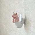 A small, cute pink pig figurine sitting on a miniature white toilet mounted on a bathroom wall.