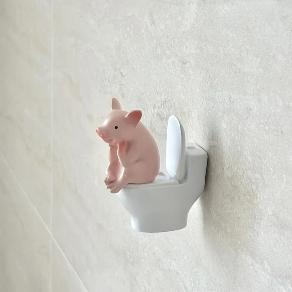 A small, cute pink pig figurine sitting on a miniature white toilet mounted on a bathroom wall.