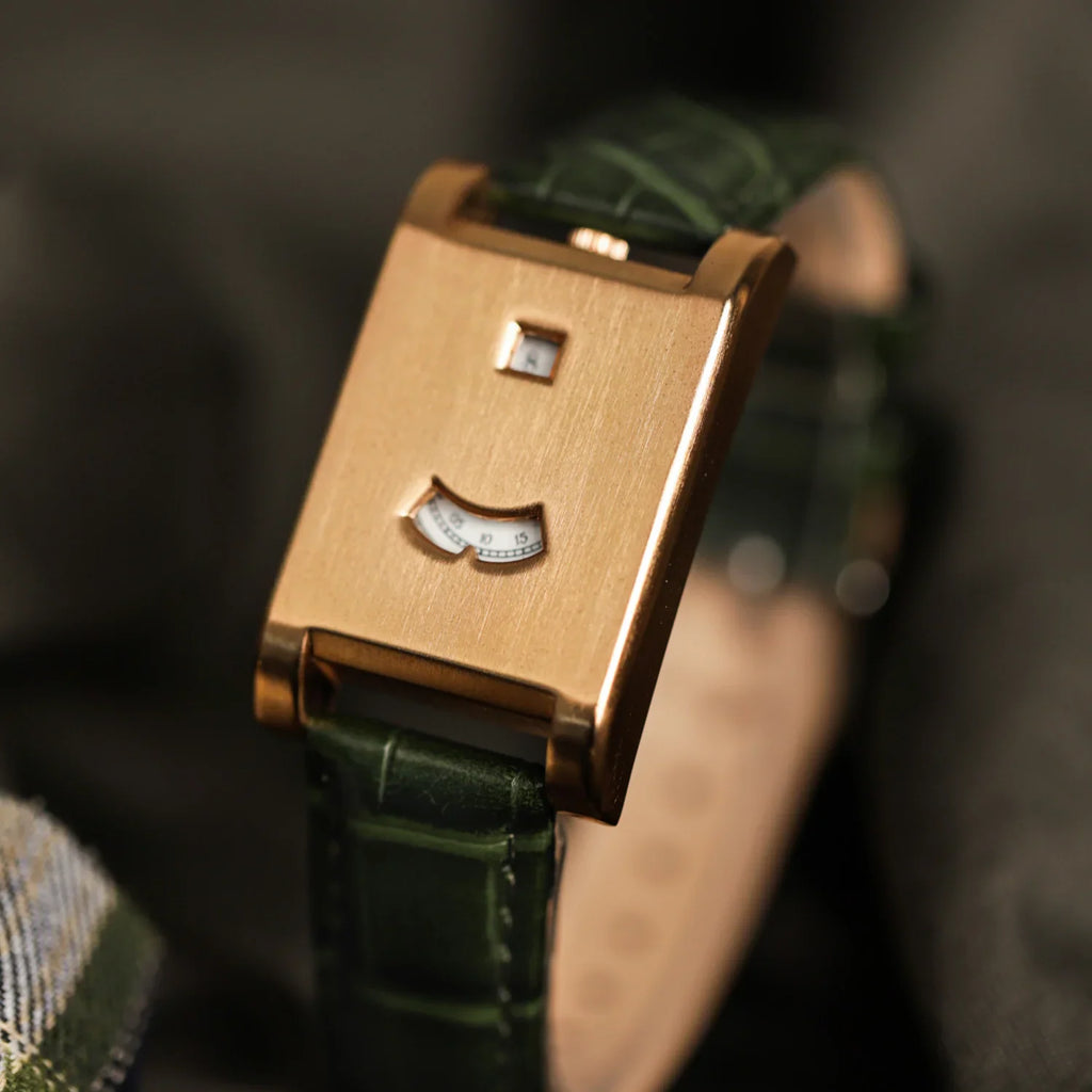 A rose gold rectangular watch with a green leather strap on a textured fabric background.