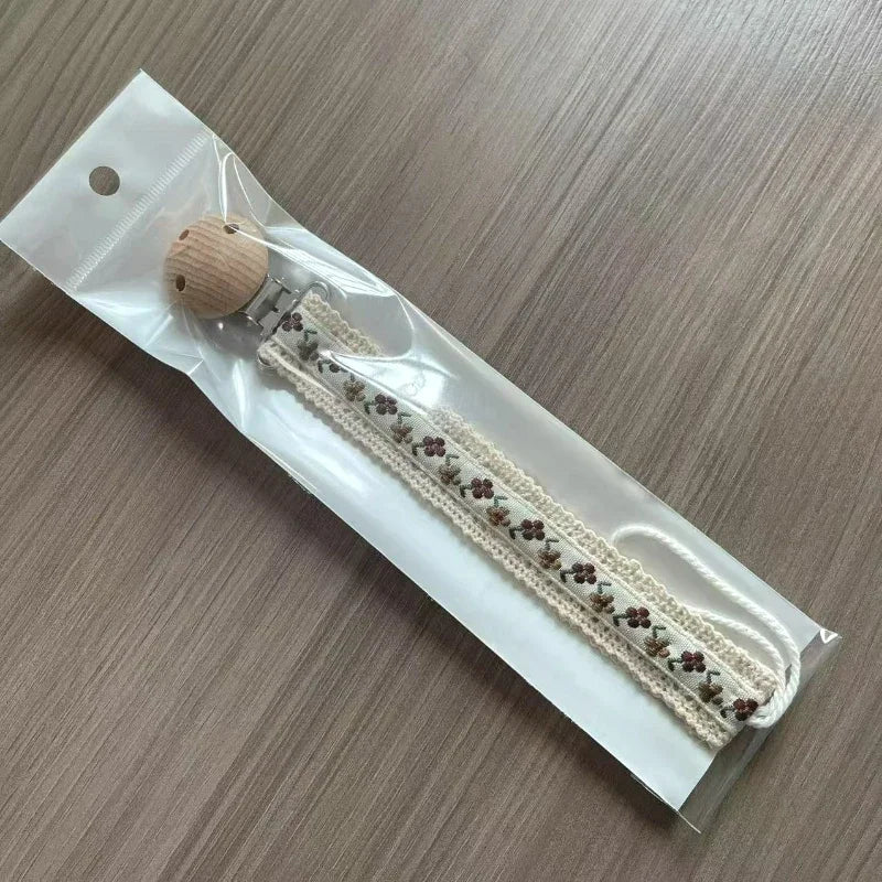 A fabric pacifier chain with a floral pattern and a wooden clip inside a plastic packaging bag.
