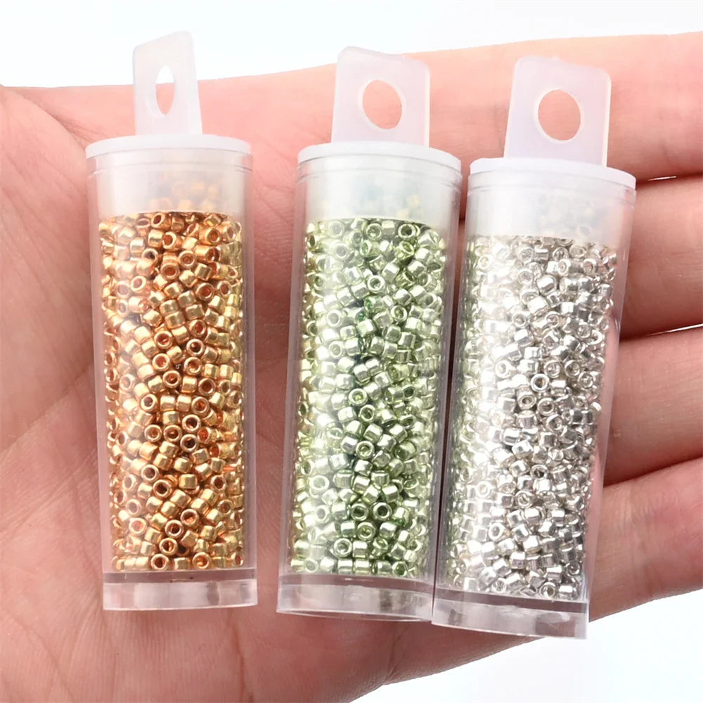 A hand holding three clear plastic tubes filled with gold, green, and silver metallic seed beads.