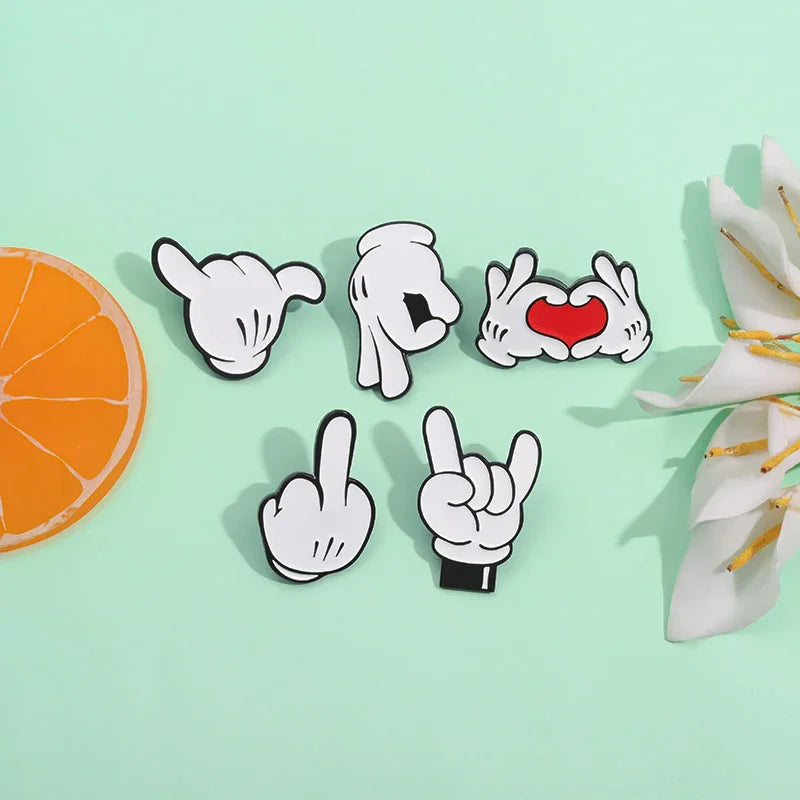 Five different cartoon hand gesture enamel pins arranged on a light green surface with an orange slice and flower