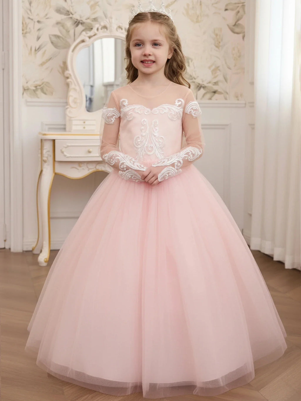 A young girl in a long, pale pink tulle dress with white lace embroidery on the bodice and long sleeves.