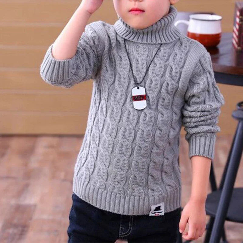 A young child wearing a grey cable-knit turtleneck sweater with a mustache logo patch.