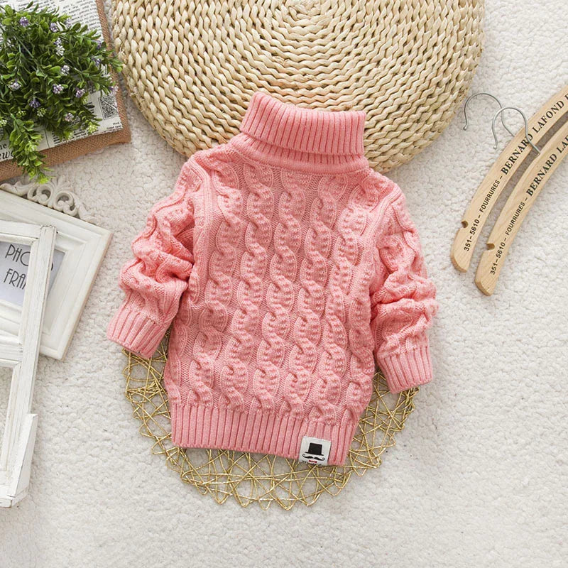 A pink cable-knit turtleneck sweater laid flat with a mustache logo patch.