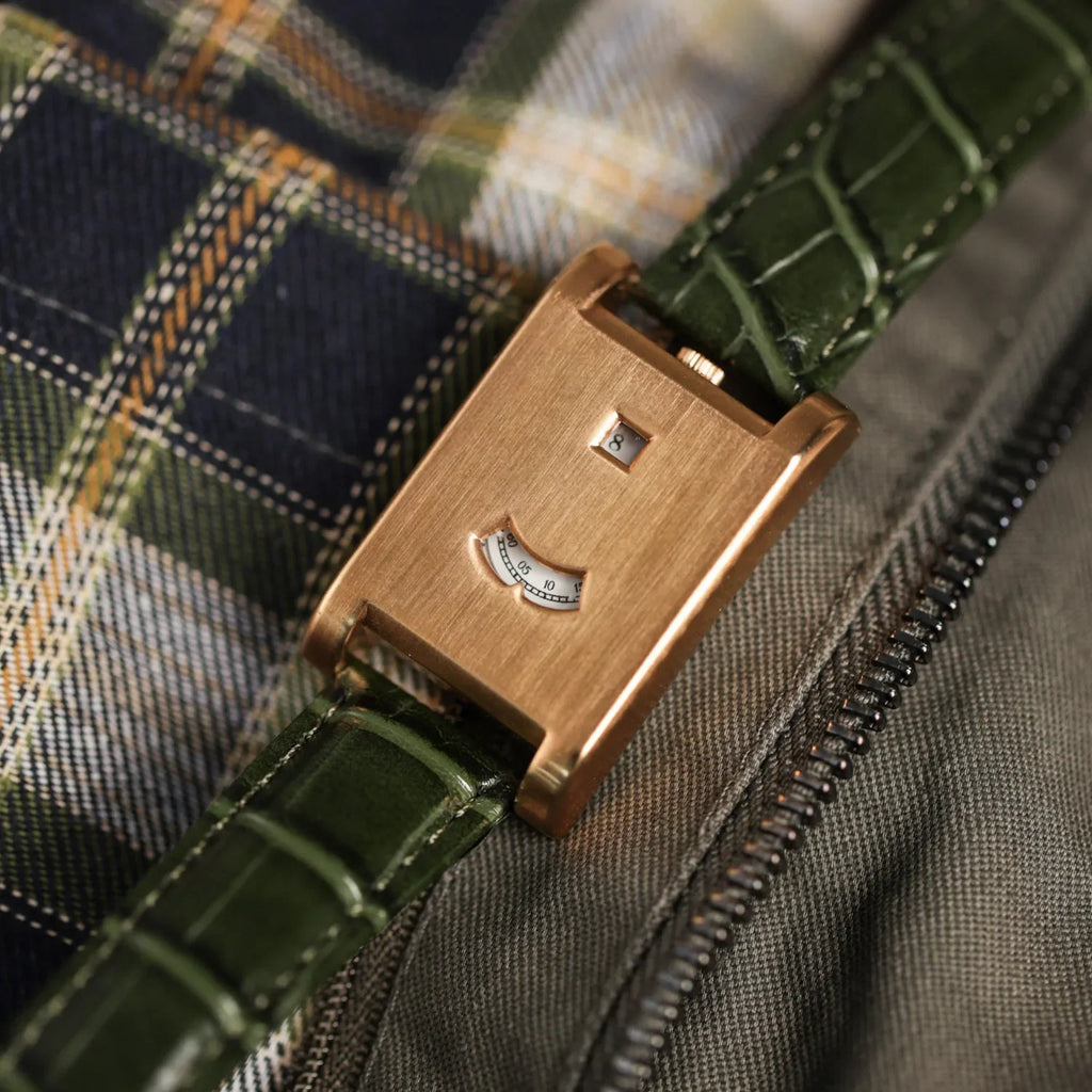 A high angle view of a rectangular rose gold watch with a green leather strap on plaid fabric.
