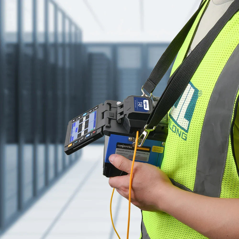 A technician wearing a high-visibility vest operating a handheld fiber optic fusion splicer