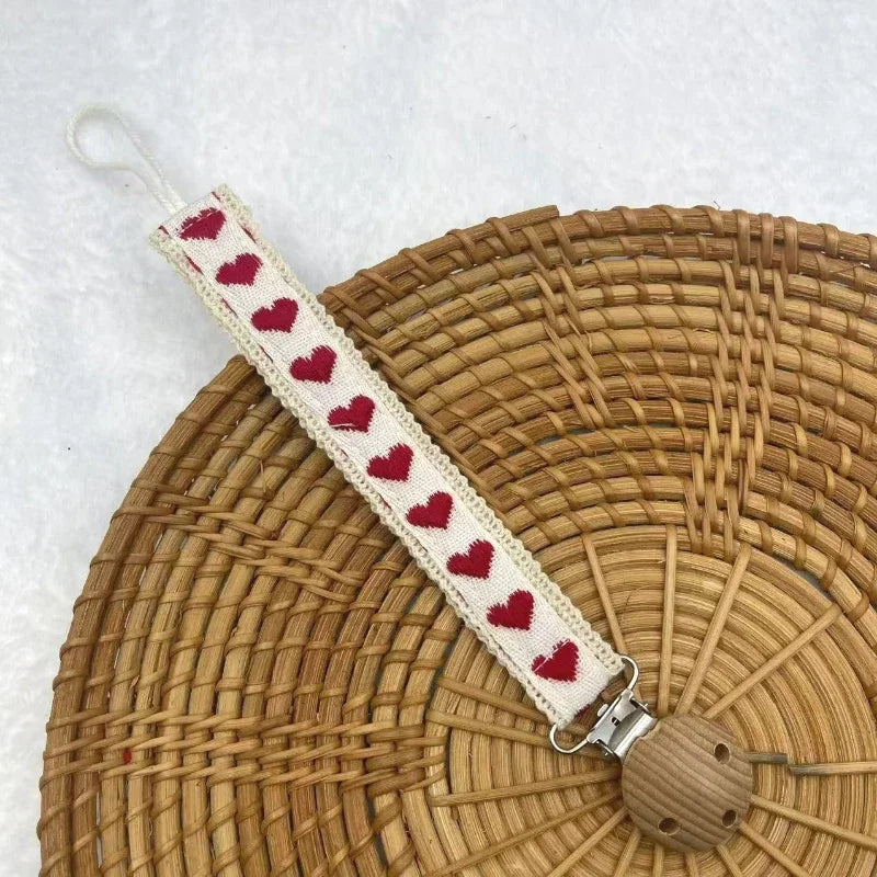 A single fabric pacifier chain with a red heart pattern and a wooden clip on a woven tray.